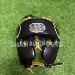 Puede incluir: Casco de boxeo negro y dorado con el texto "RIVAL PROFESSIONAL" y "SHAUZUSPORTS". El casco tiene un dise&ntilde;o acolchado con detalles en blanco y dorado. Est&aacute; dise&ntilde;ado para proteger la cabeza durante el boxeo u otros deportes de combate.