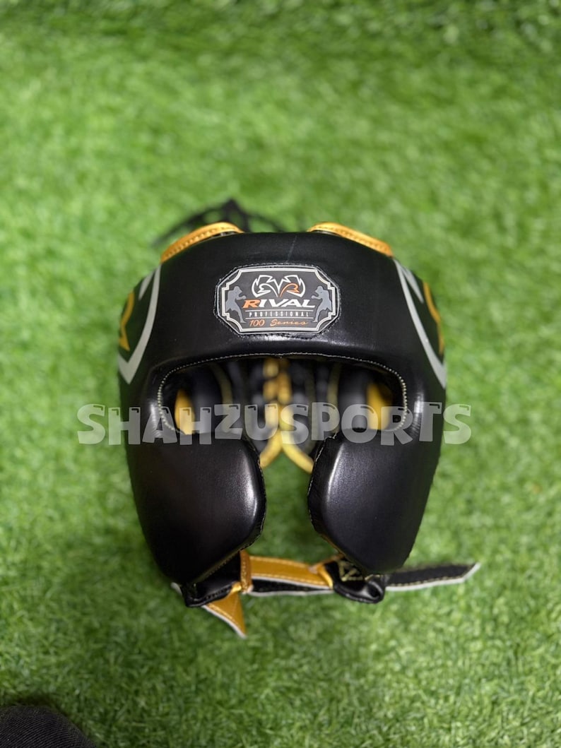 Puede incluir: Casco de boxeo negro y dorado con el texto "RIVAL PROFESSIONAL" y "SHAUZUSPORTS". El casco tiene un dise&ntilde;o acolchado con detalles en blanco y dorado. Est&aacute; dise&ntilde;ado para proteger la cabeza durante el boxeo u otros deportes de combate.