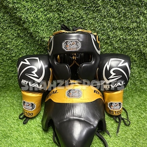 Puede incluir: Un conjunto de equipo de boxeo negro y dorado, que incluye un protector de cabeza, guantes y un protector de ingle, todos con el logotipo "Rival". Los guantes tienen un dise&ntilde;o de rayo blanco. El equipo se exhibe sobre una superficie verde y herbosa.