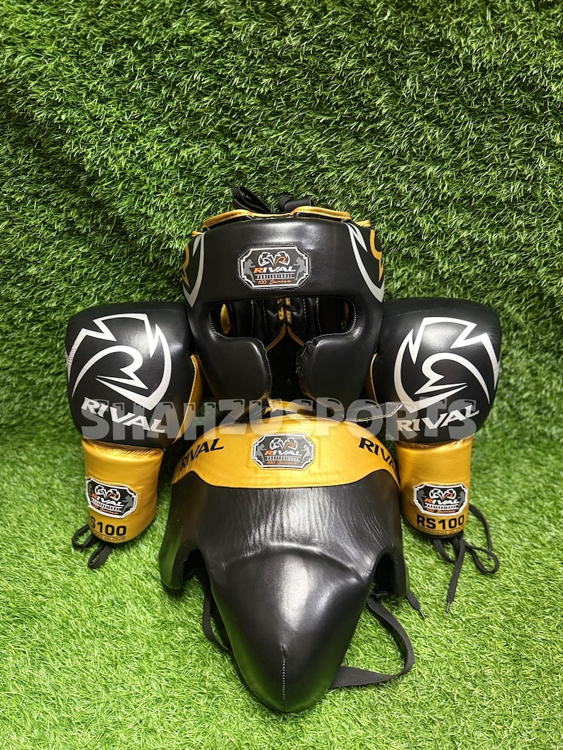 Puede incluir: Un conjunto de equipo de boxeo negro y dorado, que incluye un protector de cabeza, guantes y un protector de ingle, todos con el logotipo "Rival". Los guantes tienen un dise&ntilde;o de rayo blanco. El equipo se exhibe sobre una superficie verde y herbosa.