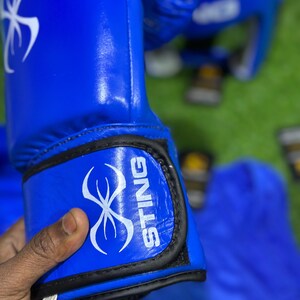May include: Bright blue boxing gloves with the word "STING" in white lettering. The gloves have a white graphic design and black trim. A matching headgear is visible in the background.