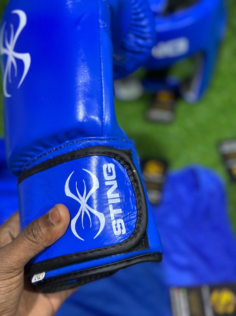 May include: Bright blue boxing gloves with the word "STING" in white lettering. The gloves have a white graphic design and black trim. A matching headgear is visible in the background.