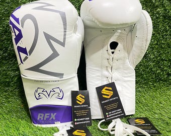 May include: Two white boxing gloves with purple and grey detailing, marked with "REAL" and "RFX GUERRERO". The gloves have an angled lace track and are shown on a green surface, accompanied by business cards.