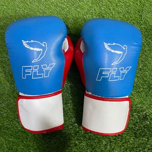 Puede incluir: Un par de guantes de boxeo azules y rojos con la palabra "FLY" impresa en blanco. Los guantes tienen una correa de muñeca blanca y un gráfico de ala. Los guantes están sobre una superficie verde.