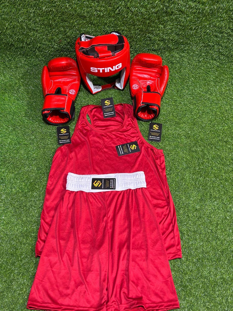 May include: A complete red boxing set, including gloves, headgear with "STING" text, a tank top, and shorts with a white waistband. The set is arranged on a green surface, ideal for boxing enthusiasts.