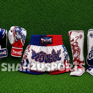 Puede incluir: Un conjunto de equipo de Muay Thai que incluye dos guantes de boxeo, un pantal&oacute;n corto y dos espinilleras. Los art&iacute;culos son blancos, rojos y azules con gr&aacute;ficos de tigre y el logo de Twins. El pantal&oacute;n tiene texto en tailand&eacute;s al frente.