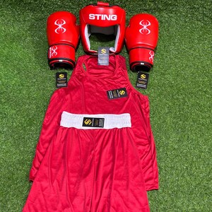 May include: A complete red boxing set, including gloves, headgear, a tank top, and shorts with a white waistband. The gloves and headgear feature the "STING" logo and a stylized design. The set is arranged on a green surface.