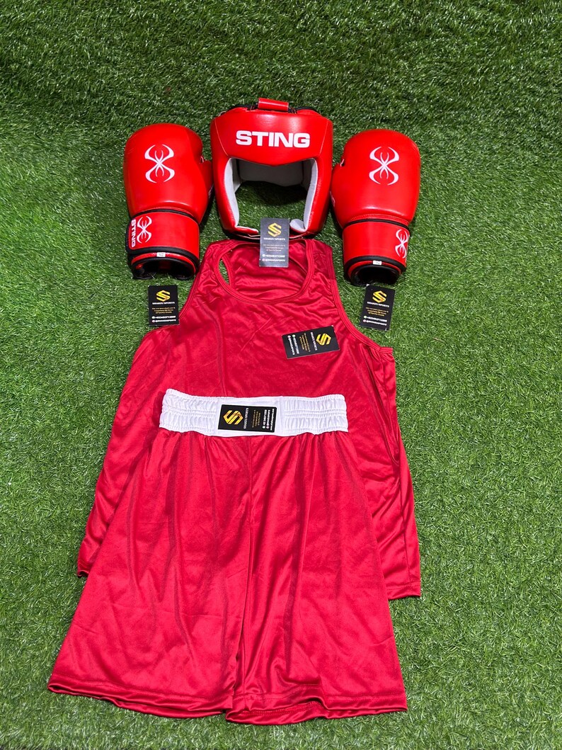 May include: A complete red boxing set, including gloves, headgear, a tank top, and shorts with a white waistband. The gloves and headgear feature the "STING" logo and a stylized design. The set is arranged on a green surface.