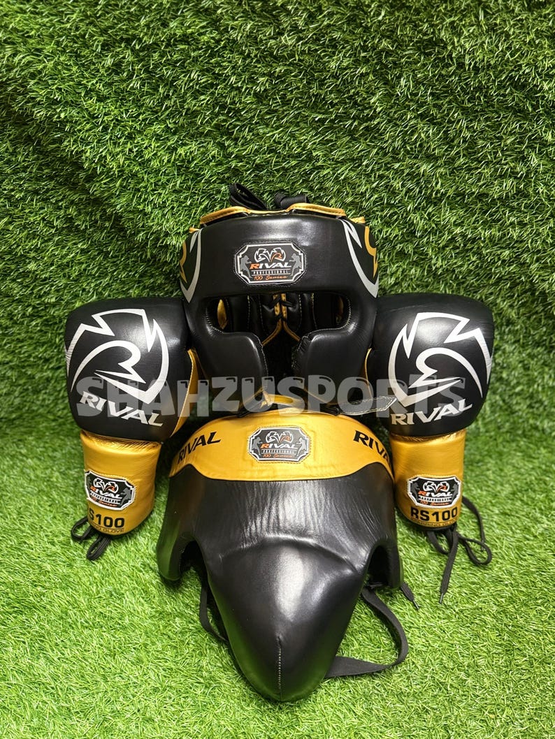 Puede incluir: Un conjunto de equipo de boxeo negro y dorado, que incluye un protector de cabeza, un protector facial y guantes de boxeo. Los guantes y el casco presentan el logotipo "RIVAL". El equipo se muestra sobre una superficie verde.
