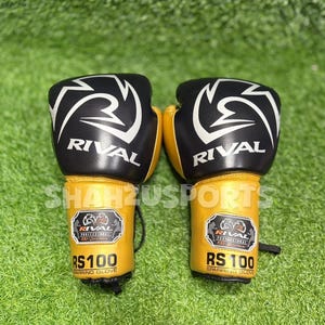 Puede incluir: Un par de guantes de boxeo negros y dorados con la palabra "RIVAL" en blanco. Los guantes tienen un dise&ntilde;o blanco en la parte superior y el texto "RS 100 SPARRING GLOVE" es visible en la zona de la mu&ntilde;eca. Sobre un fondo verde.