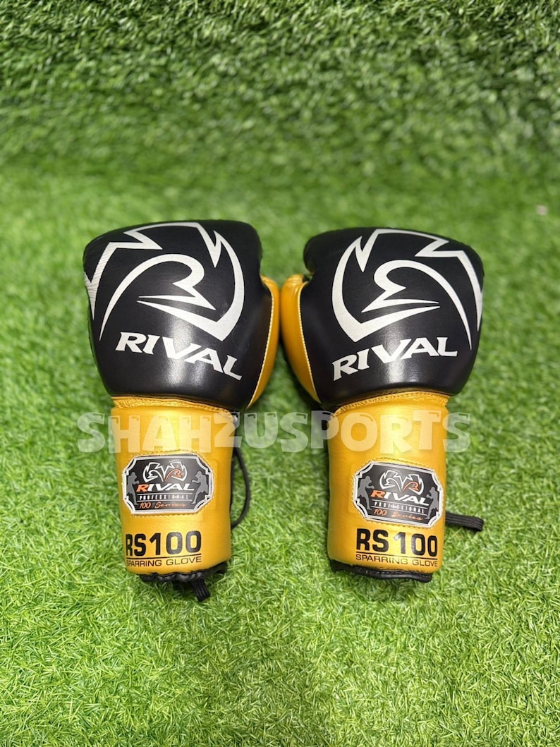 Puede incluir: Un par de guantes de boxeo negros y dorados con la palabra "RIVAL" en blanco. Los guantes tienen un dise&ntilde;o blanco en la parte superior y el texto "RS 100 SPARRING GLOVE" es visible en la zona de la mu&ntilde;eca. Sobre un fondo verde.