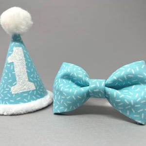May include: A blue and white bow tie and a blue party hat with a white pom-pom and the number "1" in white glitter.