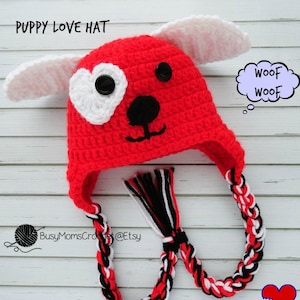 May include: Red crocheted puppy love hat with white ears, a white heart on one eye, and black button eyes. The hat has a long braided tie with red, white, and black yarn. The text "Woof Woof" is in a thought bubble.