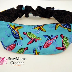 May include: A blue headband with a black elastic band and a colorful sea turtle pattern. The headband is made of fabric and has a repeating pattern of sea turtles, starfish, and bubbles. The text "BusyMoms Crochet" and "Etsy.com" is printed on the headband.