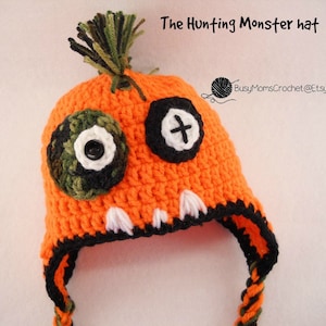 May include: Orange crocheted hat with black and white eye details and a green and black camouflage pom-pom. The hat has a black and white stitched mouth and a black and green camouflage chin strap. The hat is labeled "The Hunting Monster hat" and "BusyMomsCrochet@Etsy".