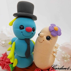Puede incluir: Una hormiga azul y amarilla con un sombrero de copa negro y una corbata negra se encuentra junto a un gusano marrón claro que lleva una flor morada. La hormiga y el gusano están sobre un tronco marrón con rosas rojas y flores blancas en el fondo.