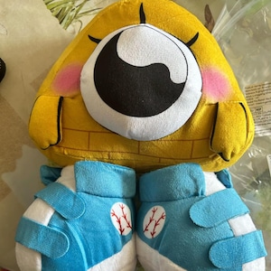 Könnte beinhalten: Ein gelbes Plüschtier mit einem großen Auge, rosa Wangen und einem ziegelartigen Muster am unteren Rand. Darunter befinden sich zwei blaue und weiße Babyschuhe mit Baseball-Nähten.