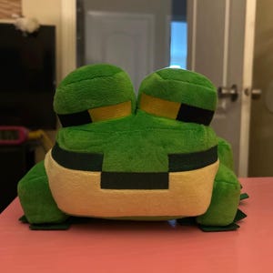 Puede incluir: Un peluche de rana de dos cabezas en tonos verdes, amarillos y crema, con un diseño pixelado. La rana está colocada sobre una superficie rosa. El diseño de la rana le da un aspecto caprichoso y de dibujos animados.