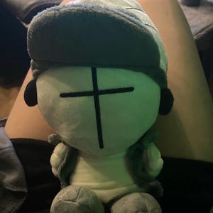 Puede incluir: Un peluche con un personaje estilizado con una cara verde claro y un dise&ntilde;o de cruz negra. El juguete tiene un sombrero y chaleco verdes a juego, con detalles negros en forma de oreja. El peluche est&aacute; sentado en el regazo de una persona.
