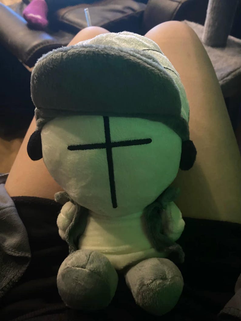 Puede incluir: Un peluche con un personaje estilizado con una cara verde claro y un dise&ntilde;o de cruz negra. El juguete tiene un sombrero y chaleco verdes a juego, con detalles negros en forma de oreja. El peluche est&aacute; sentado en el regazo de una persona.
