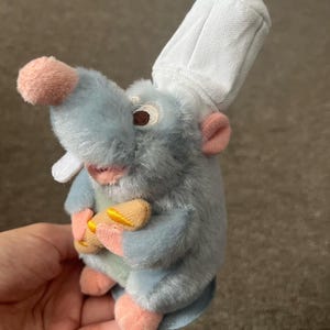 Puede incluir: Un peluche de una rata de dibujos animados con un gorro de chef blanco. La rata es azul claro con orejas, nariz y pies rosas. Sostiene dos pequeños panecillos de color canela. El juguete tiene ojos bordados y una boca sonriente.