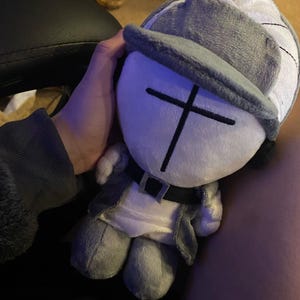 Puede incluir: Mu&ntilde;eco de peluche con gorro gris y blanco, chaqueta gris y cintur&oacute;n negro. El mu&ntilde;eco tiene una cara blanca con un dise&ntilde;o de cruz negra. El mu&ntilde;eco es un personaje de dibujos animados.