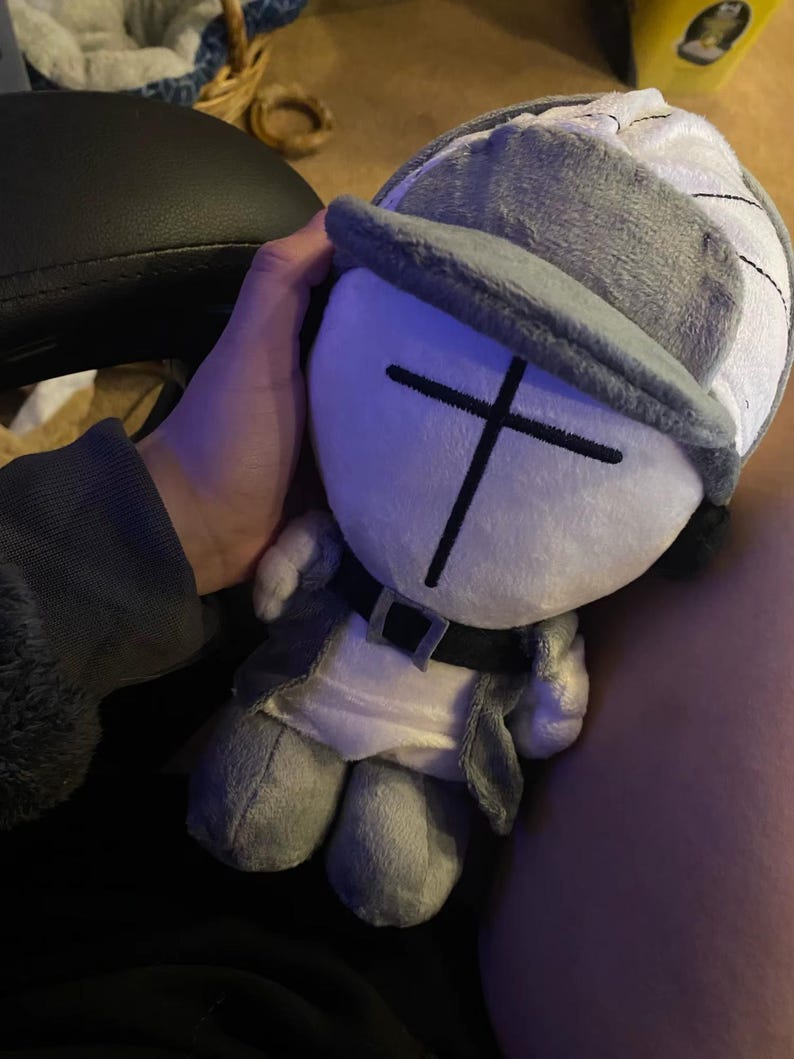 Puede incluir: Mu&ntilde;eco de peluche con gorro gris y blanco, chaqueta gris y cintur&oacute;n negro. El mu&ntilde;eco tiene una cara blanca con un dise&ntilde;o de cruz negra. El mu&ntilde;eco es un personaje de dibujos animados.
