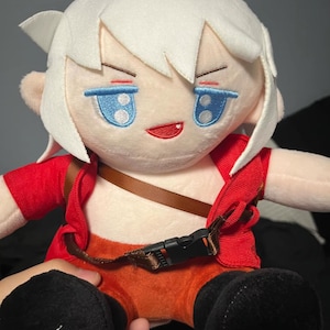 Könnte beinhalten: Ein Plüschpuppe mit weißen Haaren, blauen Augen und einem roten Outfit. Die Puppe hat einen braunen Riemen über der Brust und schwarze Stiefel. Das Gesicht der Puppe hat ein leichtes Grinsen.