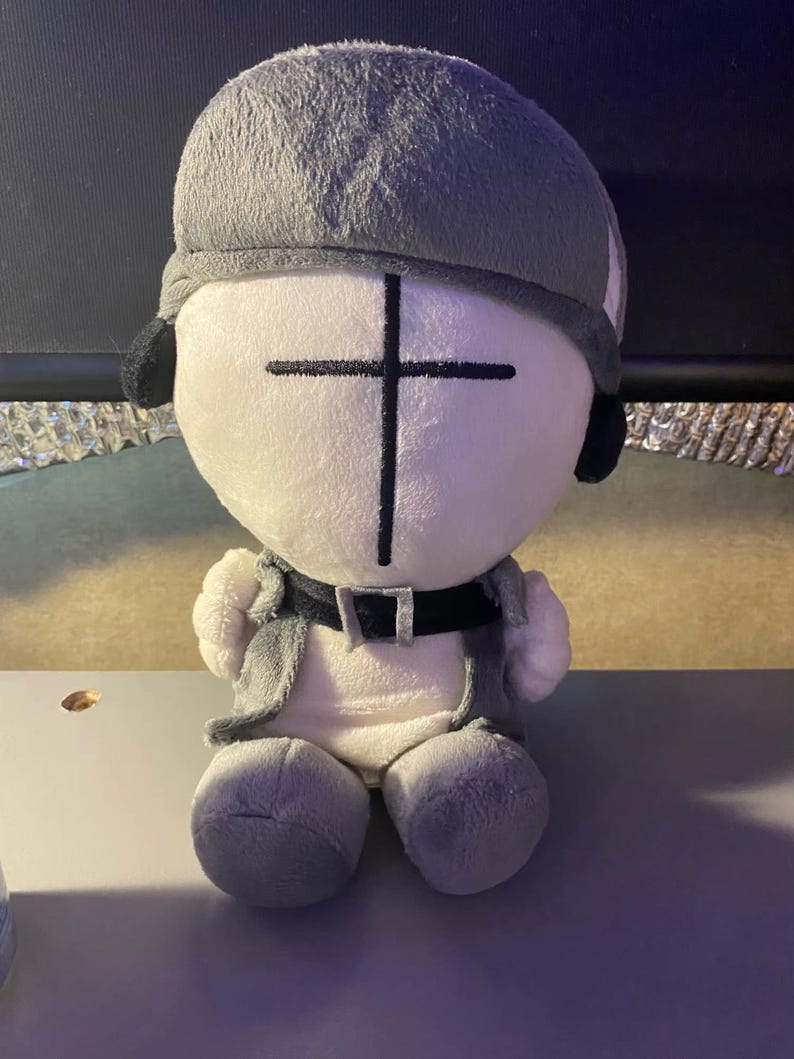 Puede incluir: Una mu&ntilde;eca de peluche con una cara blanca y un dise&ntilde;o de cruz negra. La mu&ntilde;eca tiene un casco, chaleco y pantalones grises. La mu&ntilde;eca tiene un cintur&oacute;n negro y orejeras negras. La mu&ntilde;eca est&aacute; sentada sobre una superficie blanca.