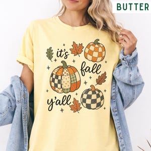 Può includere: Una t-shirt a maniche corte color giallo burro con una stampa grafica di tre zucche con motivi a scacchi, foglie autunnali e il testo "it's fall y'all" in un carattere corsivo nero, indossata sotto una giacca di jeans azzurra.
