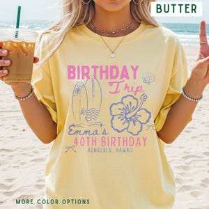 May include: Butter-colored t-shirt with a pink and blue graphic. The graphic includes the words "BIRTHDAY Trip" and "Emma's 40th Birthday Honolulu, Hawaii" with surfboards and a hibiscus flower design. The shirt is a casual, oversized fit.