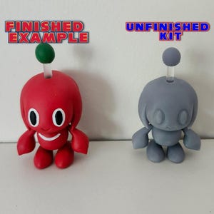 May include: A red and white character figurine with a green top, next to a grey unpainted version of the same figurine. The text "FINISHED EXAMPLE" is above the red figurine and "UNFINISHED KIT" is above the grey figurine.