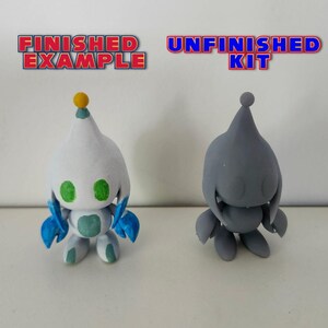May include: A finished and unfinished 3D printed figurine of a character from the Sonic the Hedgehog video game series. The finished figurine is painted white with green eyes, blue arms, and a yellow top. The unfinished figurine is grey and unpainted. The text "FINISHED EXAMPLE" is above the finished figurine and the text "UNFINISHED KIT" is above the unfinished figurine.