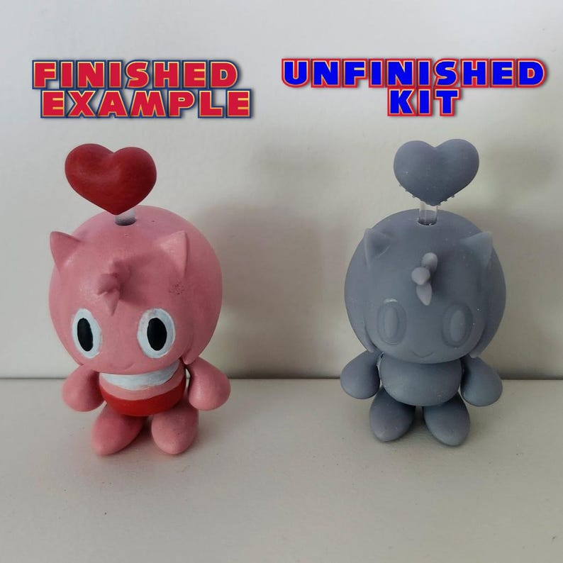 May include: A finished and unfinished pink and grey character figurine with a heart on top. The finished figurine is pink with white details and a red heart. The unfinished figurine is grey and has no paint.