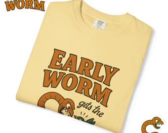 Könnte beinhalten: Hellgelbes T-Shirt mit dem Schriftzug "EARLY WORM" in braunen Buchstaben. Darunter eine Cartoon-Wurm-Illustration und der Satz "gets the". Das Shirt ist gefaltet und hat ein Etikett am Kragen.