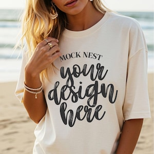 May include: A white oversized t-shirt with the text "MOCK NEST your design here" printed on the front. The shirt is being worn by a person on a beach.