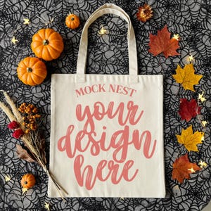May include: A cream-colored tote bag with the text "MOCK NEST your design here" in a pink script. The bag is surrounded by orange pumpkins, fall leaves, and decorative lights on a black spiderweb background.