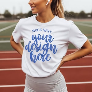 May include: White t-shirt with the text "MOCK NEST your design here" in blue script. The shirt is short-sleeved and worn by a person. The background features a red track and field surface.