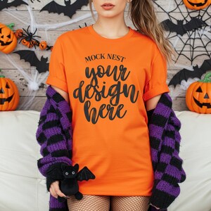 Puede incluir: Camiseta naranja con el texto "MOCK NEST your design here". Un suéter a rayas moradas y negras está sobre los hombros. Un pequeño peluche de murciélago negro se sostiene en la mano. Decoraciones temáticas de Halloween en el fondo.