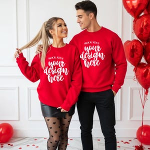 May include: Two people wearing matching red sweatshirts with the text "MOCK NEST your design here" in white script. The woman is wearing a black skirt and heart-patterned tights. Red heart-shaped balloons are in the background.