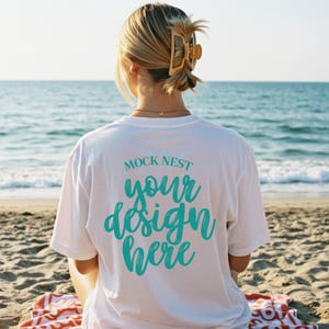 以下が含まれることがあります： 「MOCK NEST your design here」という文字がターコイズ色の筆記体で書かれた白いTシャツ。 海を背景に、ビーチに座っている人。 金色のヘアクリップをつけたブロンドの髪の人。