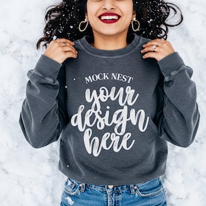 May include: A dark gray sweatshirt with the text "MOCK NEST your design here" in white script. The sweatshirt is worn with blue jeans. The image is taken in a snowy environment.