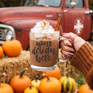 Puede incluir: Una taza de vidrio transparente llena de una bebida caliente, cubierta con crema batida y una ramita de canela. La taza tiene el texto "your design here" y "MOCK NEST" impreso. Las calabazas y las balas de heno están en el fondo.