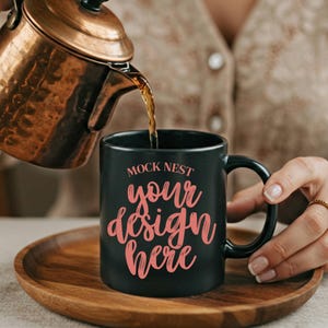 Op de afbeelding: Een zwarte koffiemok met de tekst "MOCK NEST your design here" in roze script wordt gevuld met koffie uit een koperen theepot. De mok staat op een houten dienblad.