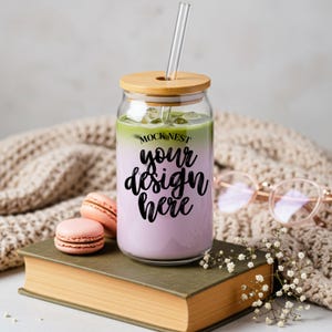 Pode incluir: Um copo de vidro transparente com tampa de bambu e canudo de vidro, preenchido com uma bebida em camadas. A bebida tem um topo verde e um fundo lavanda, com o texto "MOCK NEST your design here". Dois macarons rosa e um livro estão em primeiro plano.