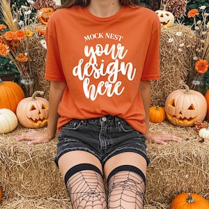 Puede incluir: Camiseta naranja con el texto "MOCK NEST your design here" en blanco. La persona lleva pantalones cortos de mezclilla negros y medias de red negras. Las calabazas y las balas de heno están en el fondo, creando un tema de otoño o Halloween.