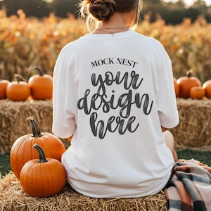Puede incluir: Camiseta blanca con el texto "MOCK NEST your design here" en una fuente de escritura. La camiseta se usa al aire libre con calabazas y balas de heno en el fondo, creando un tema de cosecha de otoño.