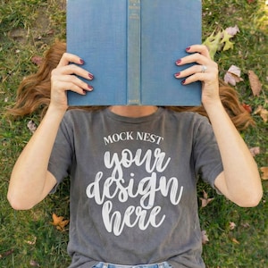 May include: A person lying on grass, holding a blue book in front of their face. They are wearing a gray t-shirt with the text "MOCK NEST your design here" in white. The person is also wearing blue denim shorts.