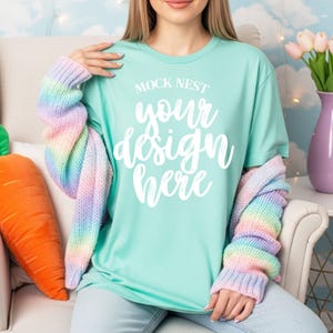 May include: A light teal t-shirt with the text "MOCK NEST your design here" in white script. A pastel rainbow cardigan is draped over the shoulders. A large orange carrot pillow is on the left.