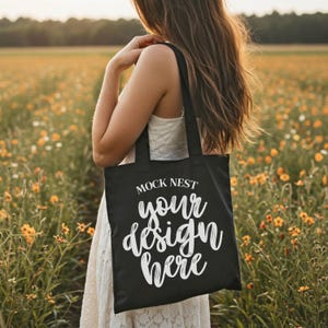Könnte beinhalten: Schwarze Tragetasche mit dem Text "MOCK NEST your design here" in weißer Schrift. Die Tasche wird von einer Person in einem Feld mit gelben Blumen getragen. Die Tasche hat zwei schwarze Griffe.